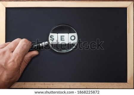 SEO, Search Engine Optimization concept.  A cropped hand holding a magnifying glass magnifies the word SEO on block letters on black background 