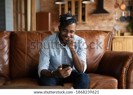 Smiling young african ethnicity guy in eyeglasses looking at smartphone screen, enjoying communicating distantly in dating application or reading email with pleasant news, sitting on sofa at home. Royalty-Free Stock Photo #1868239621
