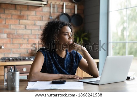 Thoughtful young mixed race african woman looking in distance, thinking of financial problems, feeling confused of overspending money while calculating monthly budget or paying bills at home. Royalty-Free Stock Photo #1868241538