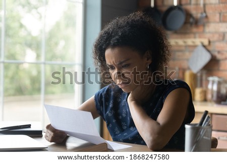Frustrated young african american woman reading paper letter with bad news, stressed of getting bank debt or loan rejection notification, feeling confused of termination notice, sitting at table. Royalty-Free Stock Photo #1868247580