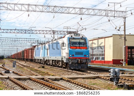 A beautiful new electric locomotive BKG1 with freight wagons stands in front of a traffic light at a train station, awaiting departure. Autumn photo. Royalty-Free Stock Photo #1868256235