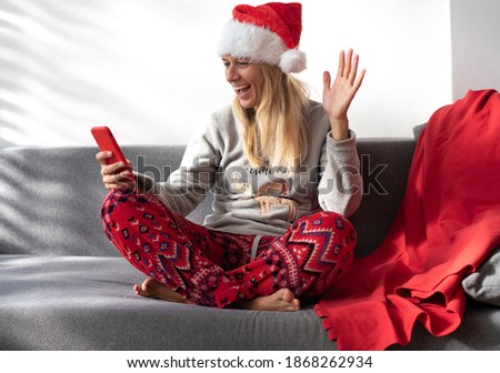 Woman sit on grey sofa in Christmas pyjamas and funny cap, with phone and talking with friends online. On quarantine time.Blond female smile and very happy to get gifts for new year.