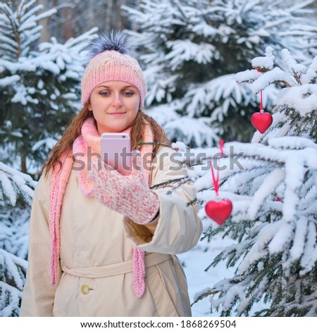 Cute woman look at phone next to the Christmas tree. Decorating trees with toys in nature and online communication with respect to social distance