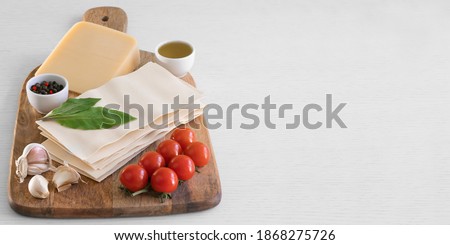 Background with ingredients for cooking Italian lasagna on wood cutting board stock photo. Ingredients: lasagna sheets, basil, cherry tomatoes, parmesan, garlic, pepper, oil, wood cutting board.