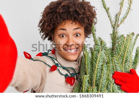Tradition of buying Christmas tree. Lovely cheerful woman with curly hair looks gladfully at camera makes photo of herself happy after winter shopping buys holiday attributes. Xmas decorations