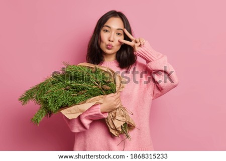 Horizontal shot of good looking brunette woman keeps lips folded and wants to kiss someone makes peace gesture tilts head holds handmade Christmas bouquet enjoys winter time. Waiting for New Year