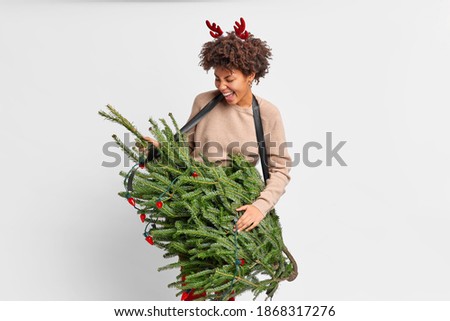 People and fun concept. Crazy mad dark skinned woman imitates playing guitar on christmas fir tree foolishes around at home on New Year Eve wears reindeer horns isolated on white background.