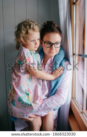 picture of self isolated lovely young caucasian female with pretty face, short dark hair, big eyes sits on the bright table with child  on her hands and looks out the window on quarantine.
