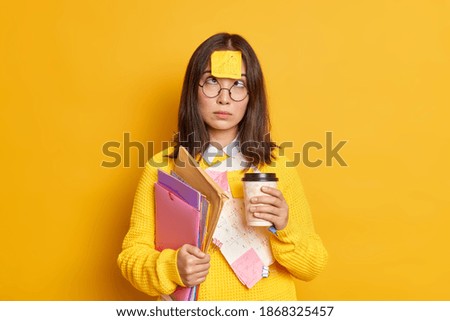 Funny female nerdy student crosses eyes has sticky note stuck on forehead has coffee break while preparing for exam much work to do holds folders and papers isolated over yellow studio background