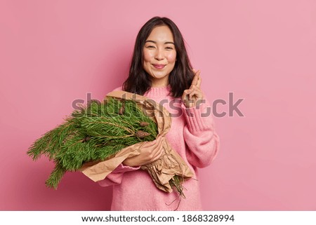 Positive brunette woman with eastern appearance holds christmas green fir tree branches arranged in bouquet makes korean like sign wears sweater poses against rosy studio wall. Happy holidays
