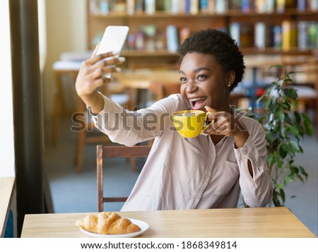 Excited black woman with coffee and smartphone taking selfie while having breakfast at cafe. Lovely African American lady making photo of herself, creating happy memories at restaurant