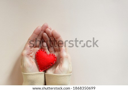 Female hands and a red felt heart with white stitches on beige background. I love you recognition. Love and romantic concept. Top view, place for text.