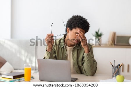 Black teenager feeling tired, rubbing irritated eyes, sitting at desk with laptop, exhausted from studying online. African American guy feeling fatigue from looking at screen, doing assignments on web Royalty-Free Stock Photo #1868354200