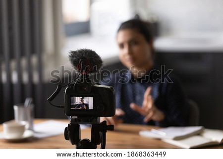 Virtual public speaking. Mixed race lady teacher trainer shooting video lecture at home office using digital camera fixed on holder. Young indian female vlogger influencer recording filming content