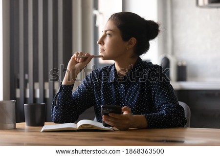 Pensive young hindu female study by desk using mobile internet distracted from making notes create new idea. Thoughtful mixed race woman looking aside of phone screen pondering planning future work Royalty-Free Stock Photo #1868363500