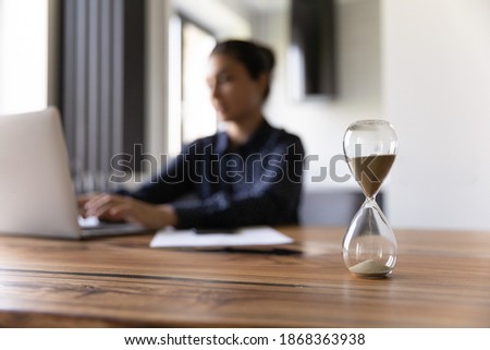 Busy young mixed race woman working studying by laptop on blurred background doing urgent job taking exam. Focus on close up sand glass posed on desk at home office. Measuring time. Deadline is coming Royalty-Free Stock Photo #1868363938