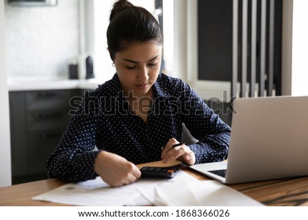 Home office. Concentrated young lady of indian ethnicity sitting at desk in modern studio apartment engaged in paperwork. Hindu woman counting preparing to pay taxes bills using computer calculator Royalty-Free Stock Photo #1868366026