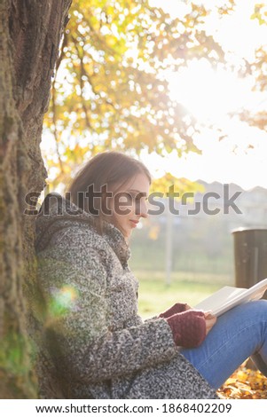 Beautiful young girl sitting and reading book under a tree in autumn.