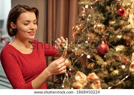 Happy young gorgeous woman in smart red dress putting decorations on Christmas tree while preparing her home for forthcoming party