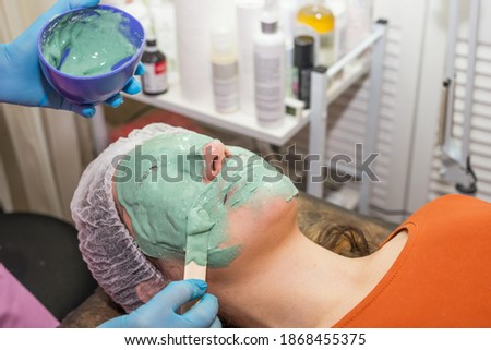 A brunette girl undergoes procedures in a spa salon, a master beautician in blue gloves applies a thick layer of a green mask, cleansing the skin of dirt, keeping an eye on her face.