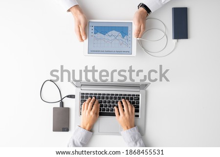 Top view of hands of two clinicians using mobile gadgets in front of one another while analyzing graphs and pressing keys of laptop keypad