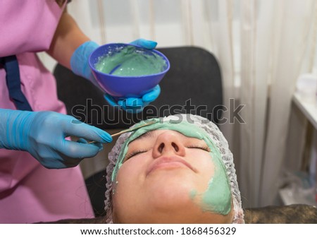 A brunette girl undergoes procedures in a spa salon, a master beautician in blue gloves applies a thick layer of a green mask, cleansing the skin of dirt, keeping an eye on her face.