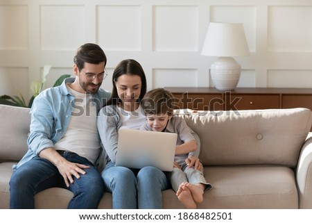 Young Caucasian family with little preschooler son relax on couch at home watch video on laptop together. Happy parents with small boy child use modern computer, rest on sofa in living room on weekend