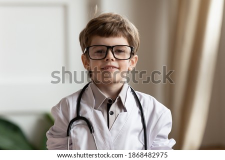 Headshot portrait of cute little boy patient in white medical uniform glasses stethoscope act as doctor in hospital. Profile picture of small child have fun play dream of pediatrician or GP career.
