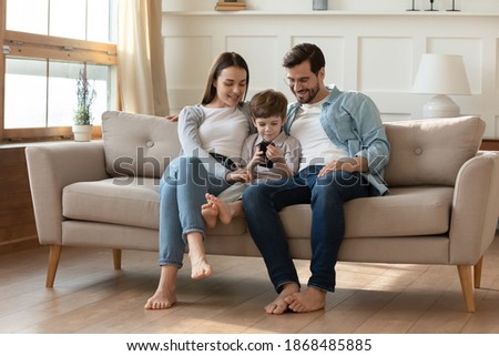 Happy young Caucasian family with small son sit relax on sofa in living room using smartphone together. Smiling parents rest on couch at home with little boy child, watch video on cellphone.