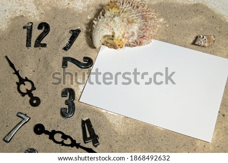digits of a clock lie in the sand, a seashell and a piece of paper nearby