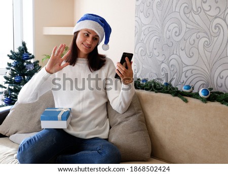 Online Christmas greetings. A woman with a phone at home in a Santa hat congratulates friends.