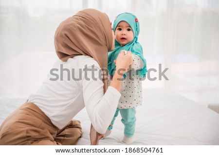Happy muslim mother and adorable little baby daughter in hijab on bed in white bedroom at home. Muslim mom hands holding baby for practice standing on bed at indoors. Love of mother and baby concept.