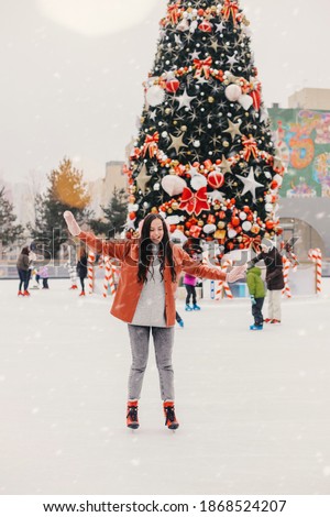 Young happy beautiful woman in stylish jacket and earmuffs learn to skate on outdoor ice rink. Cheerful girl have fun and practice skating on open ice skating rink. Winter holidays, Christmas vacation