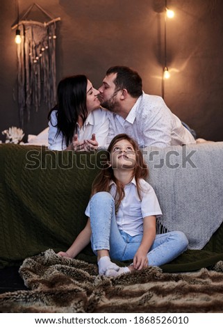 Mother and father lying on bed, kissing, and daughter sitting on the floor, looking at them in cozy dark room. Christmas and new year celebration with family. Happy winter holidays fun leisure time.