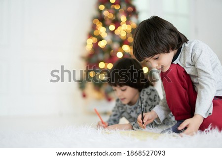 Home is the heart of holidays. Two little latin boys, twins drawing pictures with pencils while sitting on the floor at home decorated for Christmas. Siblings involved in creative activity together