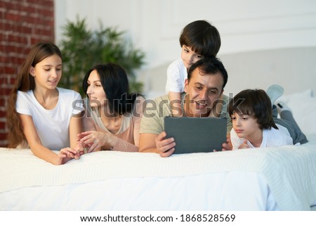 Entertainment. Happy latin family with cute little kids using digital tablet, lying on the bed together. Father looking at the screen while having video call in the morning