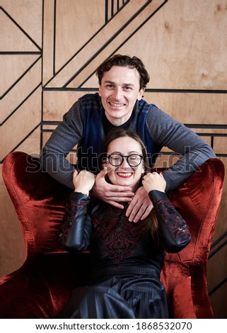Young woman in eyeglasses and man sitting on red velvet Santa armchair in front of wooden wall, smiling, laughing. Romantic relationship. Close-up picture of young couple in fancy clothes.