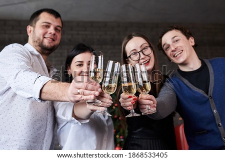 New year party celebration with friends and family at home. Close-up picture of four young people, wearing casual clothes, holding champagne glasses in dark room, smiling, laughing, doing cheers.