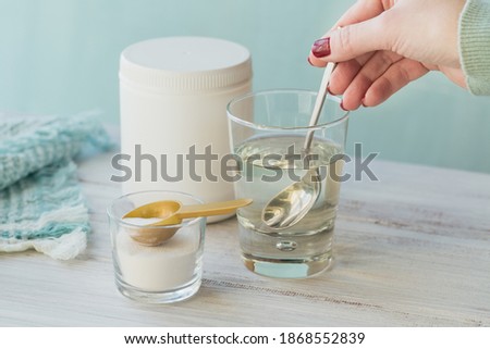 Glass with collagen dissolved in water and collagen protein powder on white wooden table. Woman's hand holds a spoon. Healthy lifestyle concept.