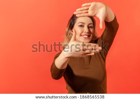 Portrait of woman with blonde hair looking through frame of fingers gesturing photo, zooming focusing to camera, observing with smile. Indoor studio shot isolated on red background