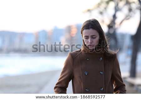 Front view portrait of a sad woman walking alone towards camera looking down on the beach in winter Royalty-Free Stock Photo #1868592577