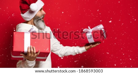 man in a Santa hat with a surprised and embarrassed expression looks at the gifts he is holding in his hands. A man does not know which gift to choose. snowing