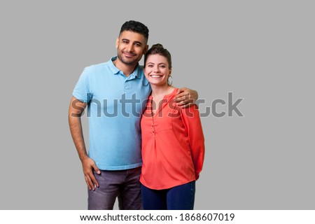 Portrait of young attractive family couple in casual wear standing together, embracing and looking at camera with sincere smile, strong relations. isolated on gray background, indoor studio shot