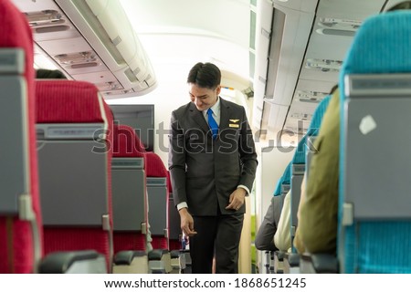 Smiling Asian male flight attendant checking the passengers on safety standard, seat belt, turn off electric devices, seat back and tray table in upright position before airplane take off and landing. Royalty-Free Stock Photo #1868651245