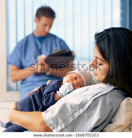 A side view of a mother holding a baby in the hospital bed while a doctor is sitting in the background checking reports Royalty-Free Stock Photo #1868705353