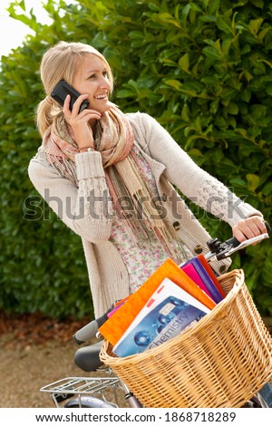 A portrait shot of a happy young blonde woman talking on a phone while standing with a bicycle and textbooks in basket.
