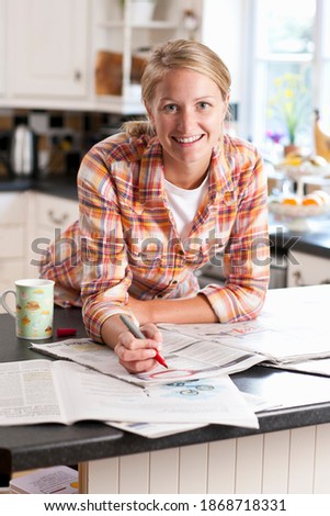 A portrait shot of a young blonde woman in checked shirt smiling at camera while circling classified ad in a newspaper.