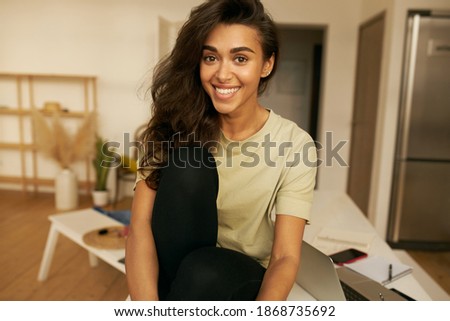 Pretty adorable young female freelancer sitting on table with open laptop, having break while working distantly from home, having relaxed joyful facial expression. Leisure and domesticity