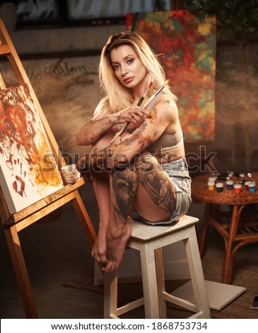 Cuddling legs seductive woman with blond hairs poses on chairs holding paintbrushes in dark atmospheric room with smoke.