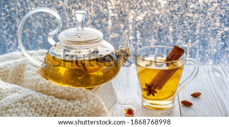 Winter warming tea with lemon and orange against the background of frosty patterns. Hot drink in a glass kettle and a cup on a wooden table in the background of the window.